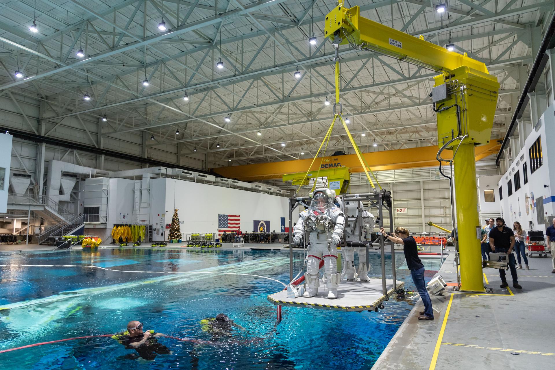 an astronaut stands on a platform about to be lowered into a large pool